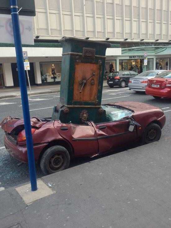 Foto gump do dia: O cofre Um enorme cofre de metal repousa sobre o teto amassado de um carro sedã preto, que está estacionado na rua. O cofre parece extremamente pesado e antigo.